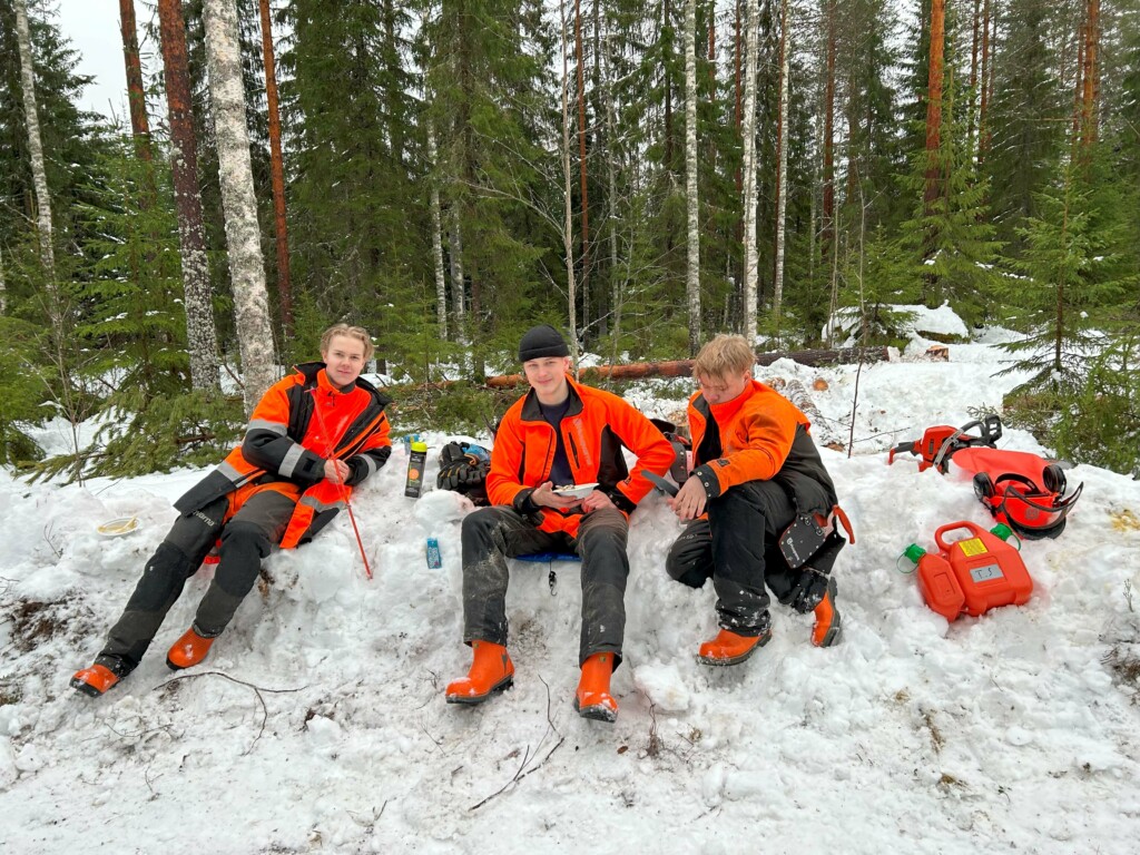 Kolme nuorta miestä istuu lumipenkassa. Taustalla metsää. Metsurin suojavaatteet päällä.