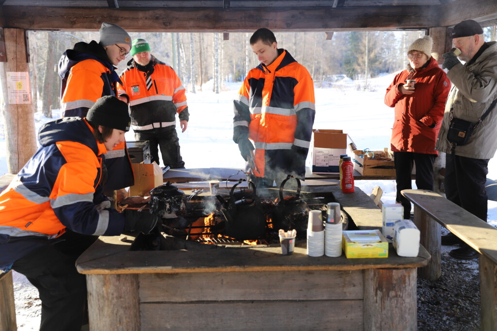 Talvi. Nuotiokatos. Kahvipannuja. Kolme metsäopiskelijaa. Kaksi vanhempaa ihmistä juo kahvia seisten. Ohjaaja seisoo taustalla.