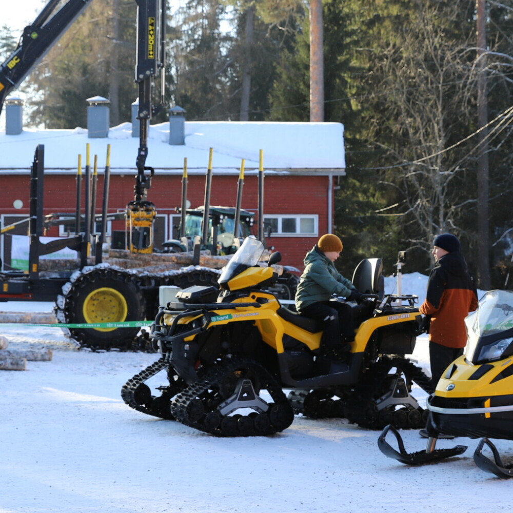 Talvinen pihamaa. Metsäkone. Moottorikelkkoja. Lapsi kelkan päällä ja pari aikuista katsomassa.