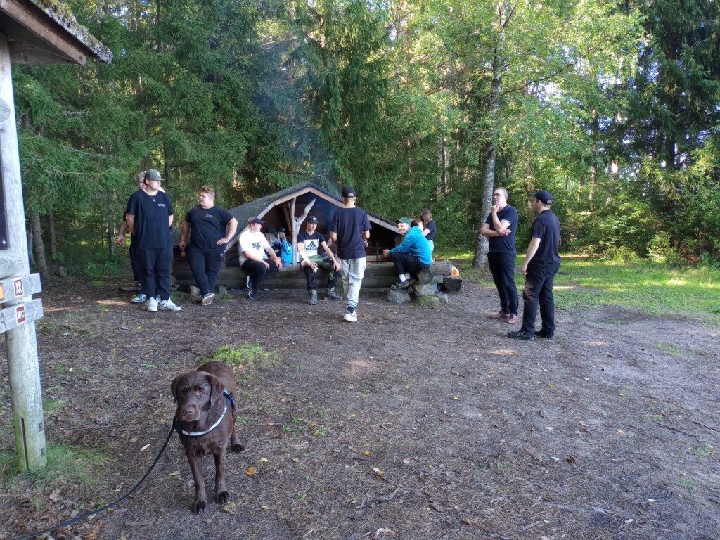 Kesä. 12 ihmistä laavun lähellä. Savu nousee laavun edestä. Kuvan etualalla seisoo märkä labradorinnoutaja. 