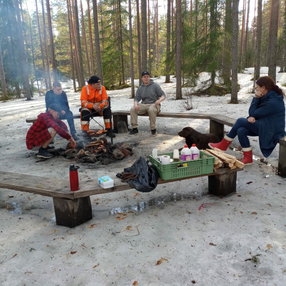 Kevättalvi. Neljä nuorta miestä ja nainen istuvat nuotiopaikalla. Retkitarvikkeita penkillä. Labradorinnoutaja makaa maassa.