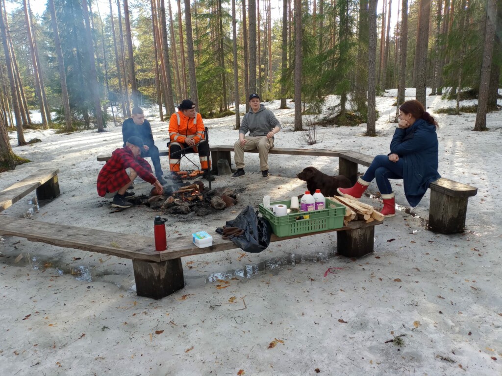 Kevättalvi. Neljä nuorta miestä ja nainen istuvat nuotiopaikalla. Retkitarvikkeita penkillä. Labradorinnoutaja makaa maassa.