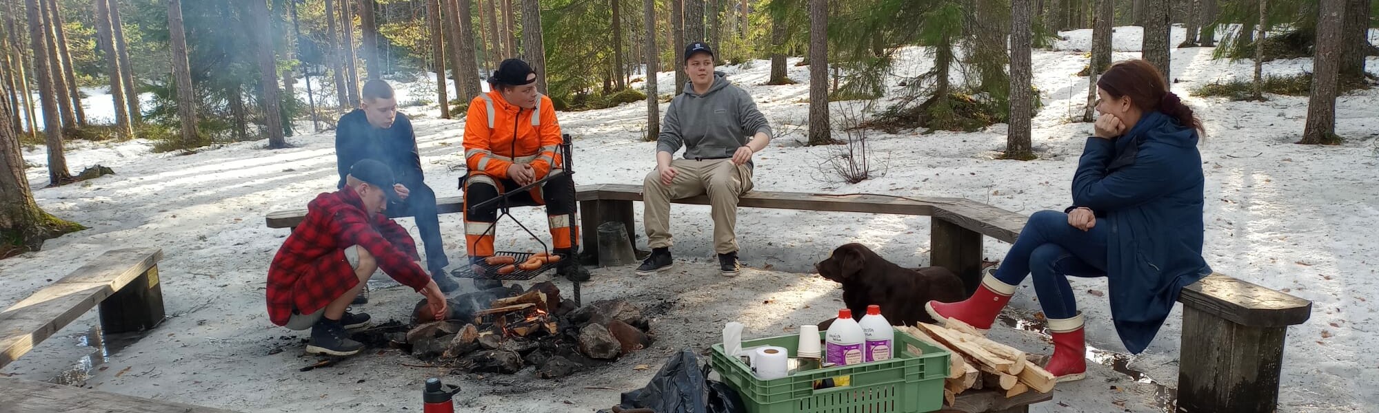 Kevättalvi. Neljä nuorta miestä ja nainen istuvat nuotiopaikalla. Retkitarvikkeita penkillä. Labradorinnoutaja makaa maassa.