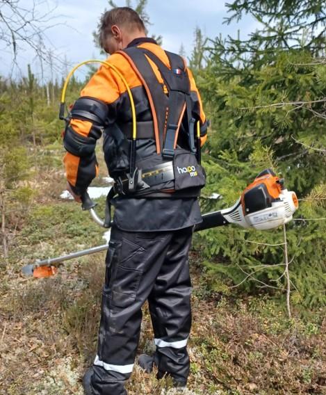Mies seisoo ulkona. Metsurin varusteet. Selässä eksoskeleton. Raivaussaha.