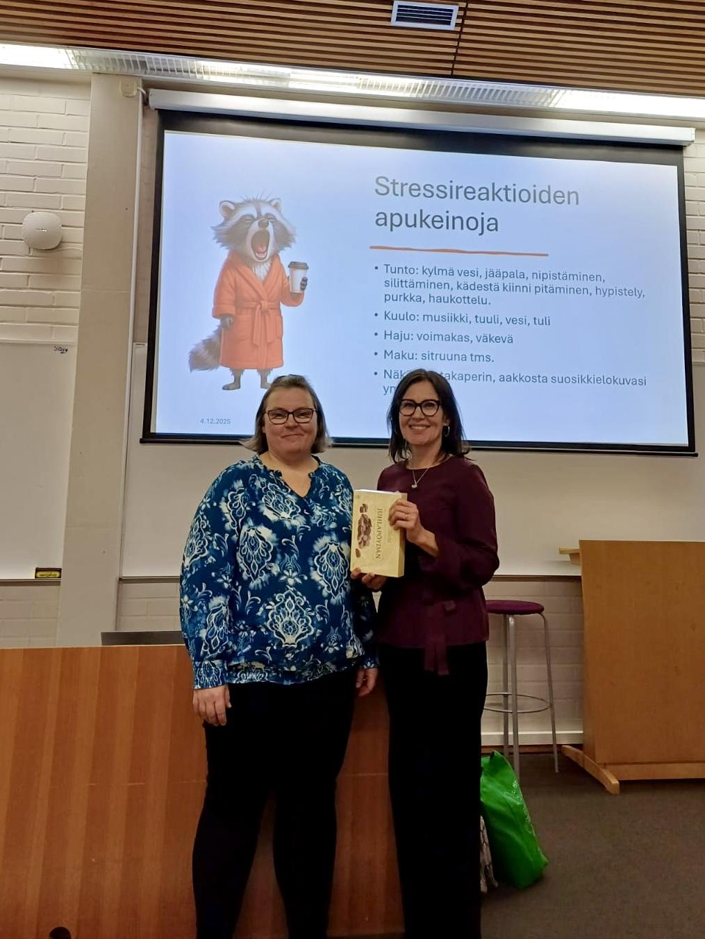 Kaksi naista seisoo vierekkäin puhujapöntön edessä ja hymyilevät kameralle. Taustalla dia, jossa otsikkona Stressireaktioiden apukeinoja.