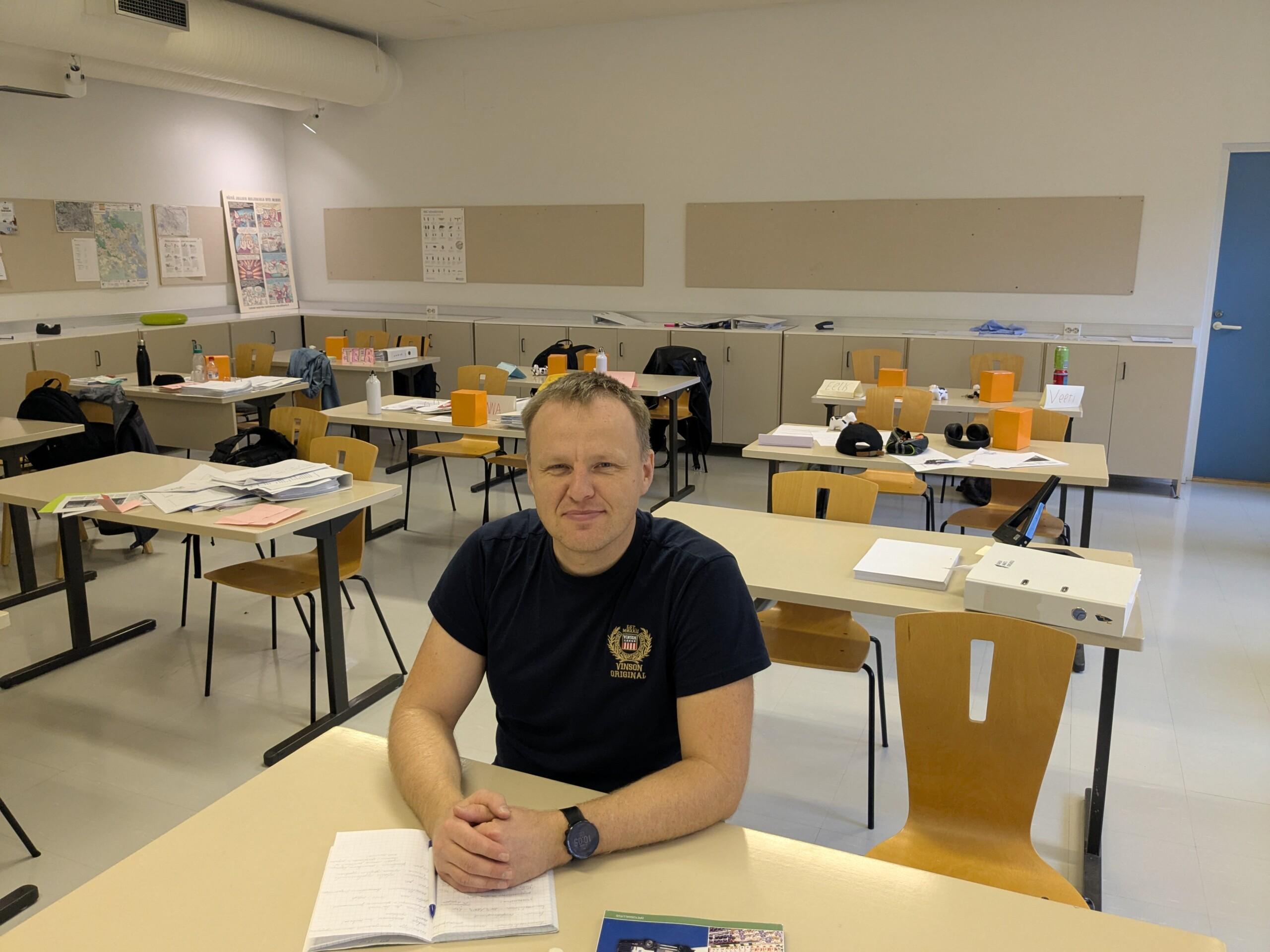 Mies istuu tyhjässä luokassa ja hymyilee kameralle.