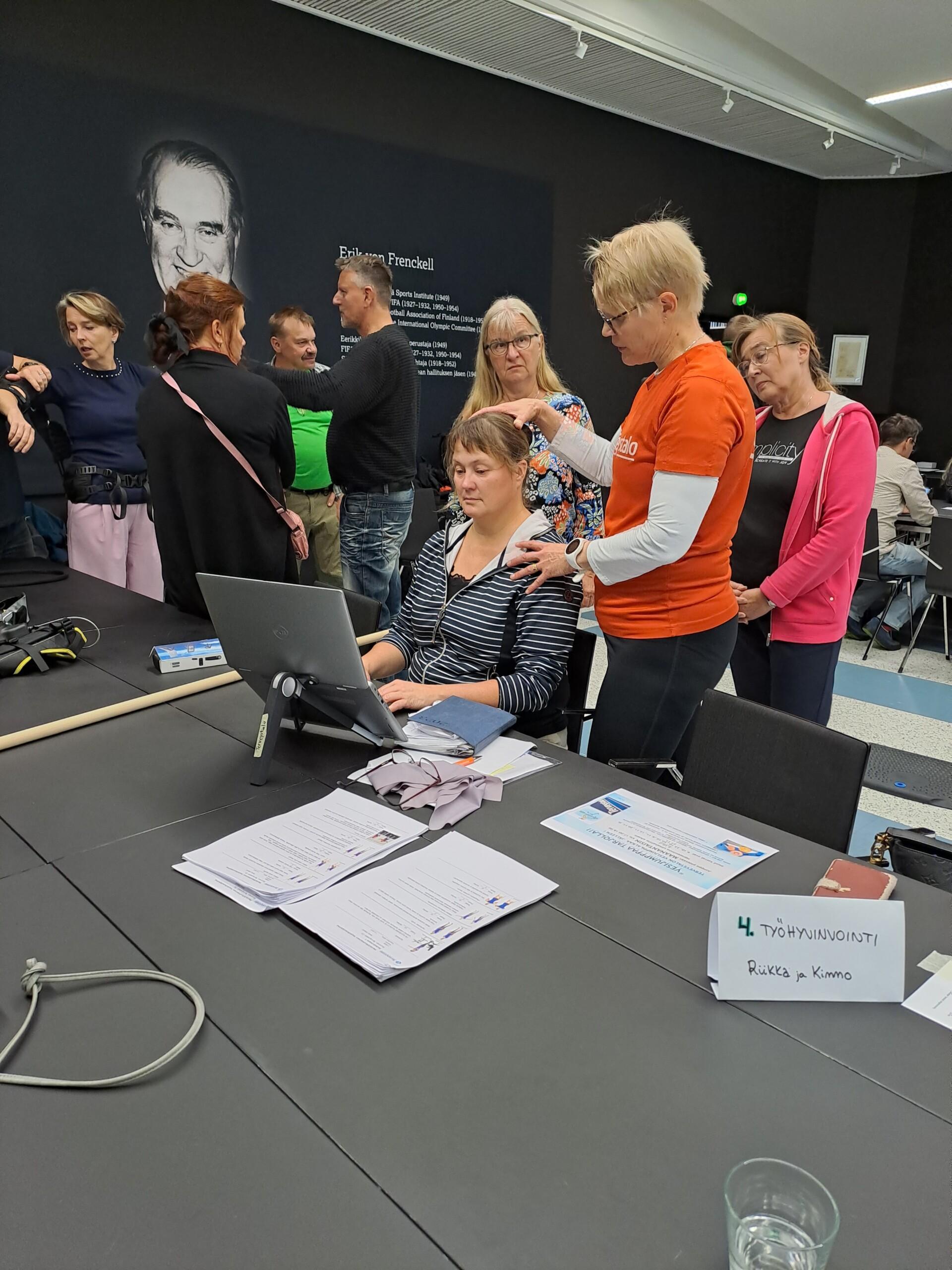Neljä naista. Yksi istuu tietokoneella. Toinen laittaa hänen päätään ja hartioitaan oikeaan asentoon. Kaksi katselee taustalla.