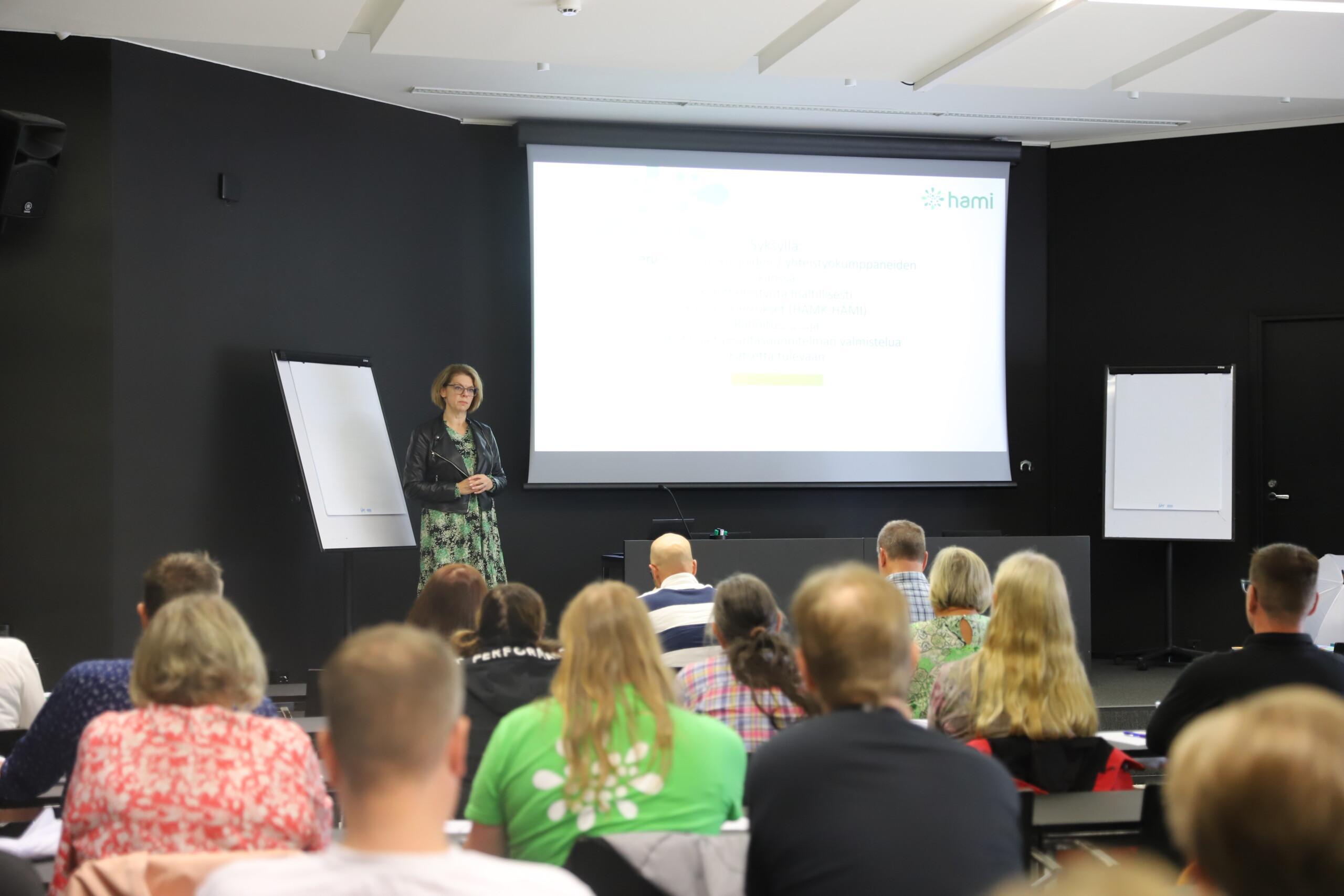 Konferenssihuone. Ihmiset kuuntelevat naispuhujaa.