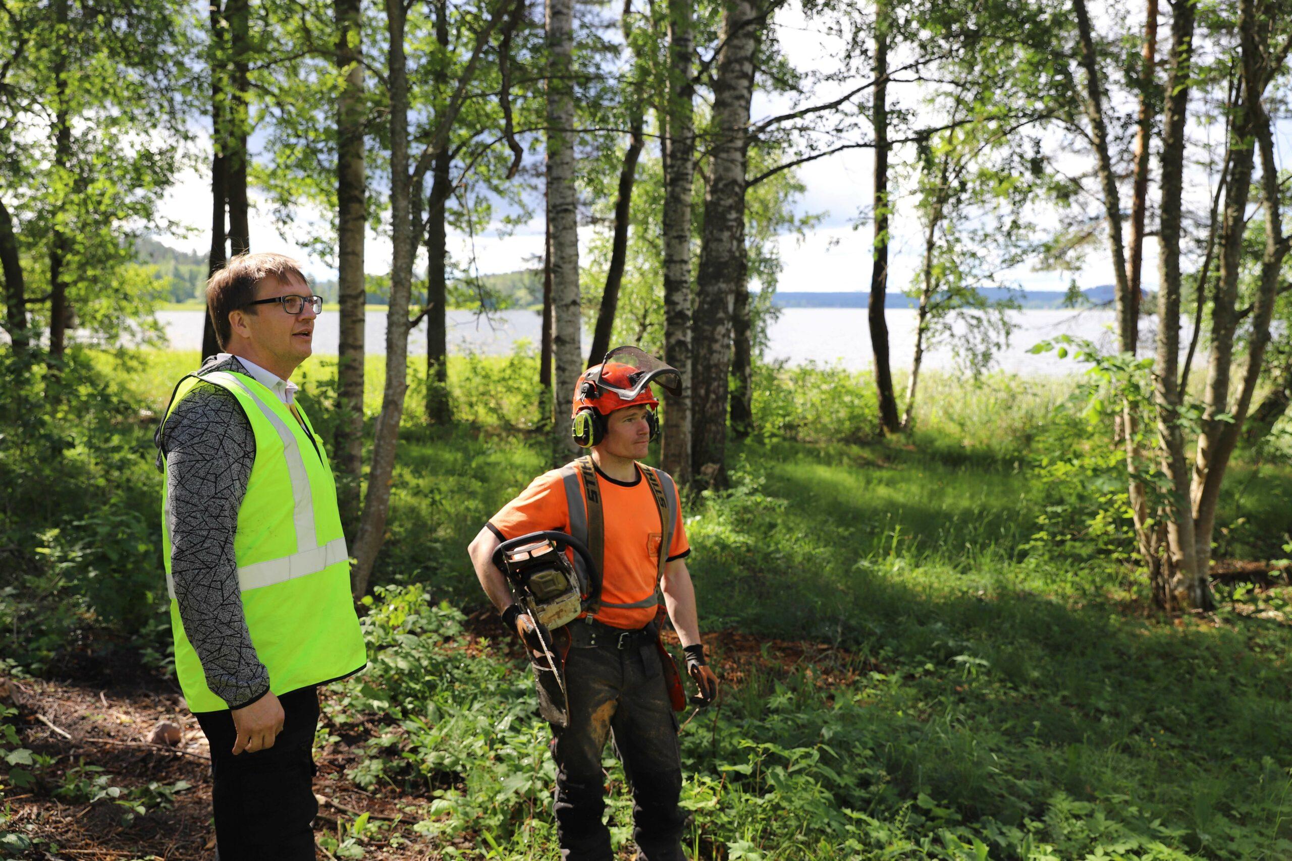 Kaksi miestä seisovat harvassa metsässä. Taustalla järvi. Toisella metsurin vaatteet, toisella turvaliivi.