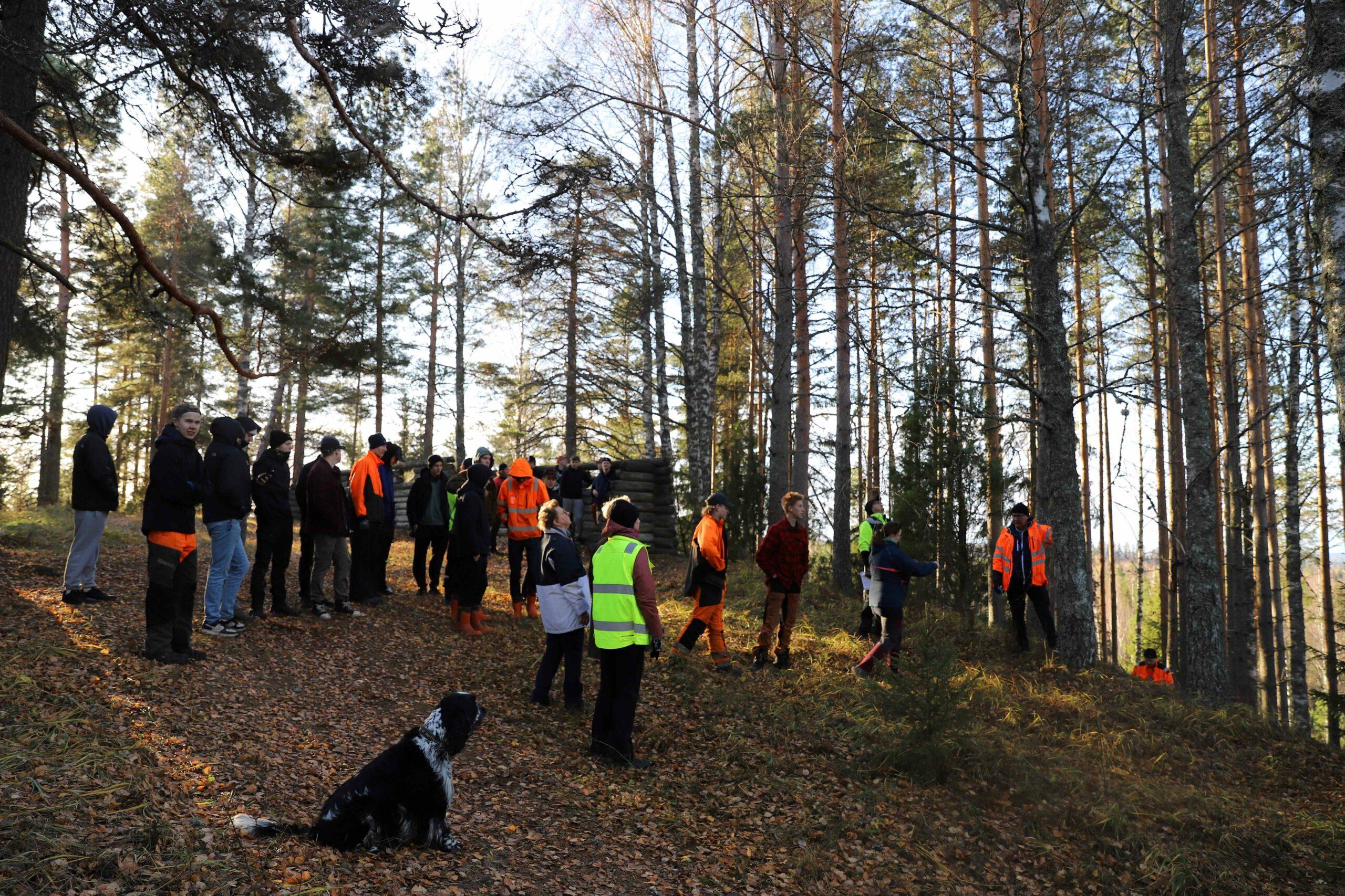Ryhmä ihmisiä seisoo ulkona rinteessä. Koira istuu etualalla. Syksyistä. Lehdettömät puut.