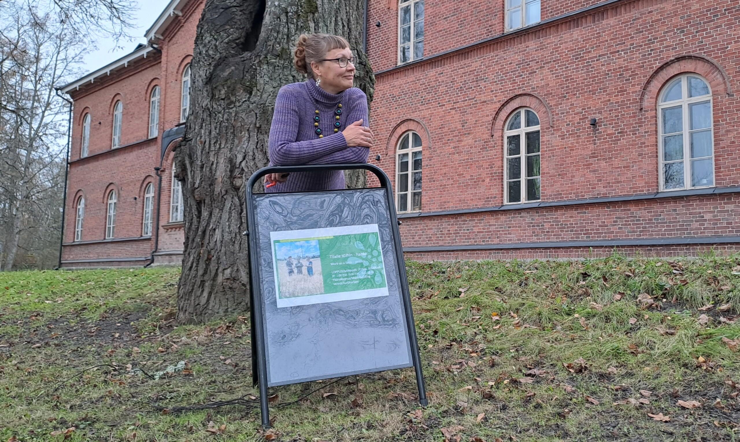 nainen nojaa ulkona mainostelineeseen. taustalla iso tiilirakennus. 