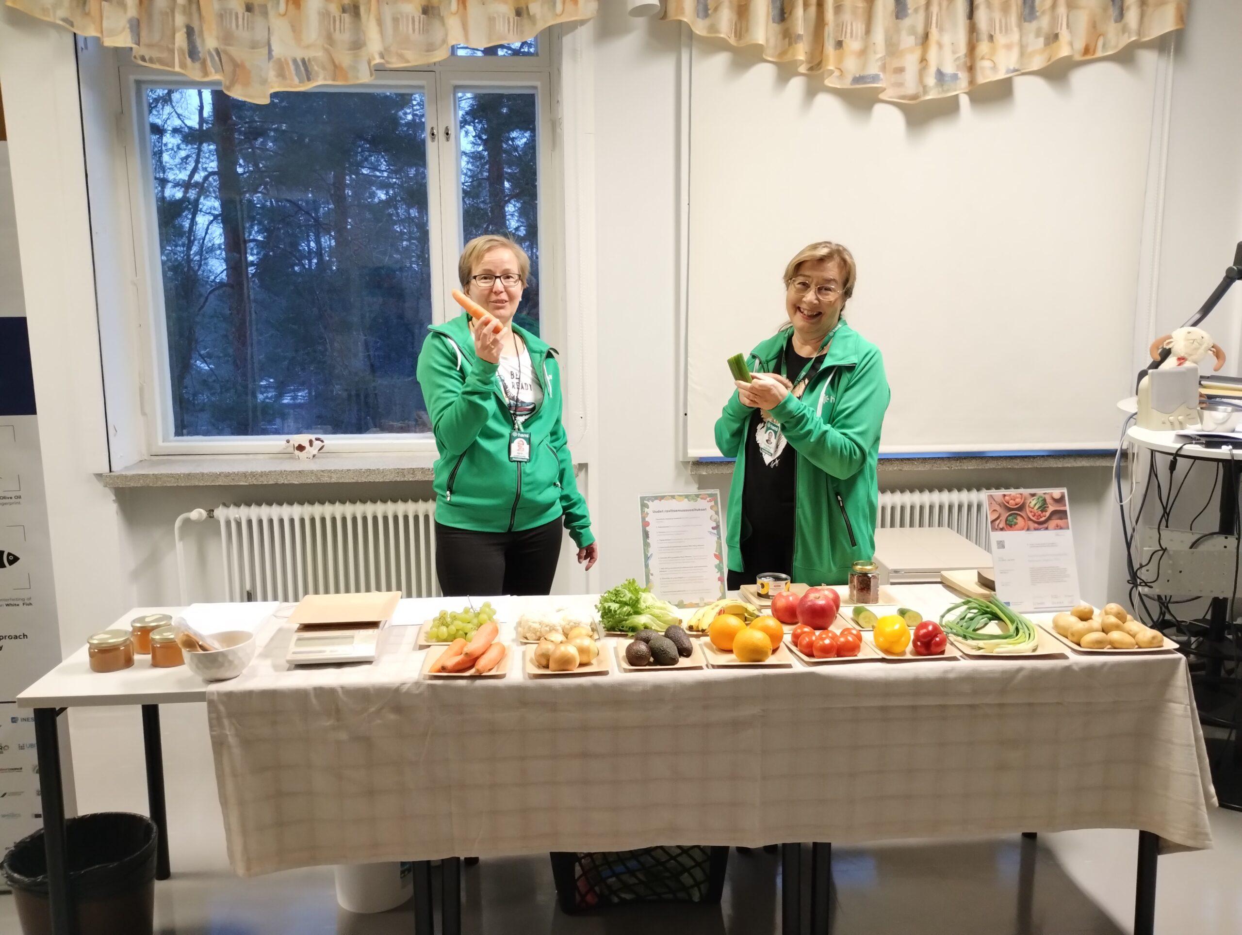 Kaksi naista seisoo esittelypöydän takana. Toisella naisella on kädessään porkkana ja toisella kurkku. Pöydällä esillä erilaisia kasviksia ja hunajaa.