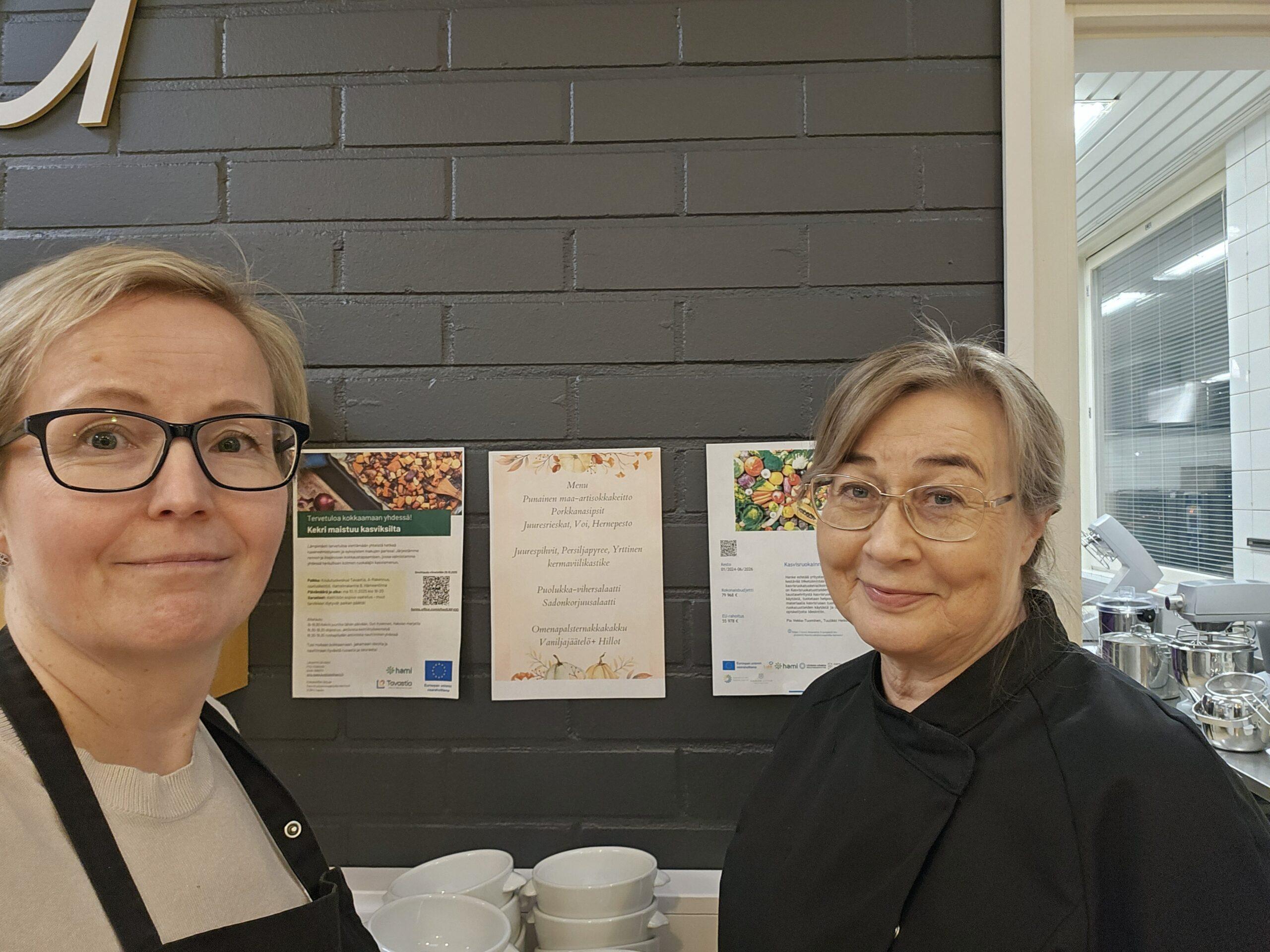 Kaksi silmälaispäistä naista katsoo kameraan hymyillen. Taustalla lautaspinoja ja julisteita seinällä.