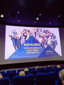 Elokuvateatteri. Valkokangas. Penkkirivistöt. Valkokankaalla otsikko "Rekryklinikka".