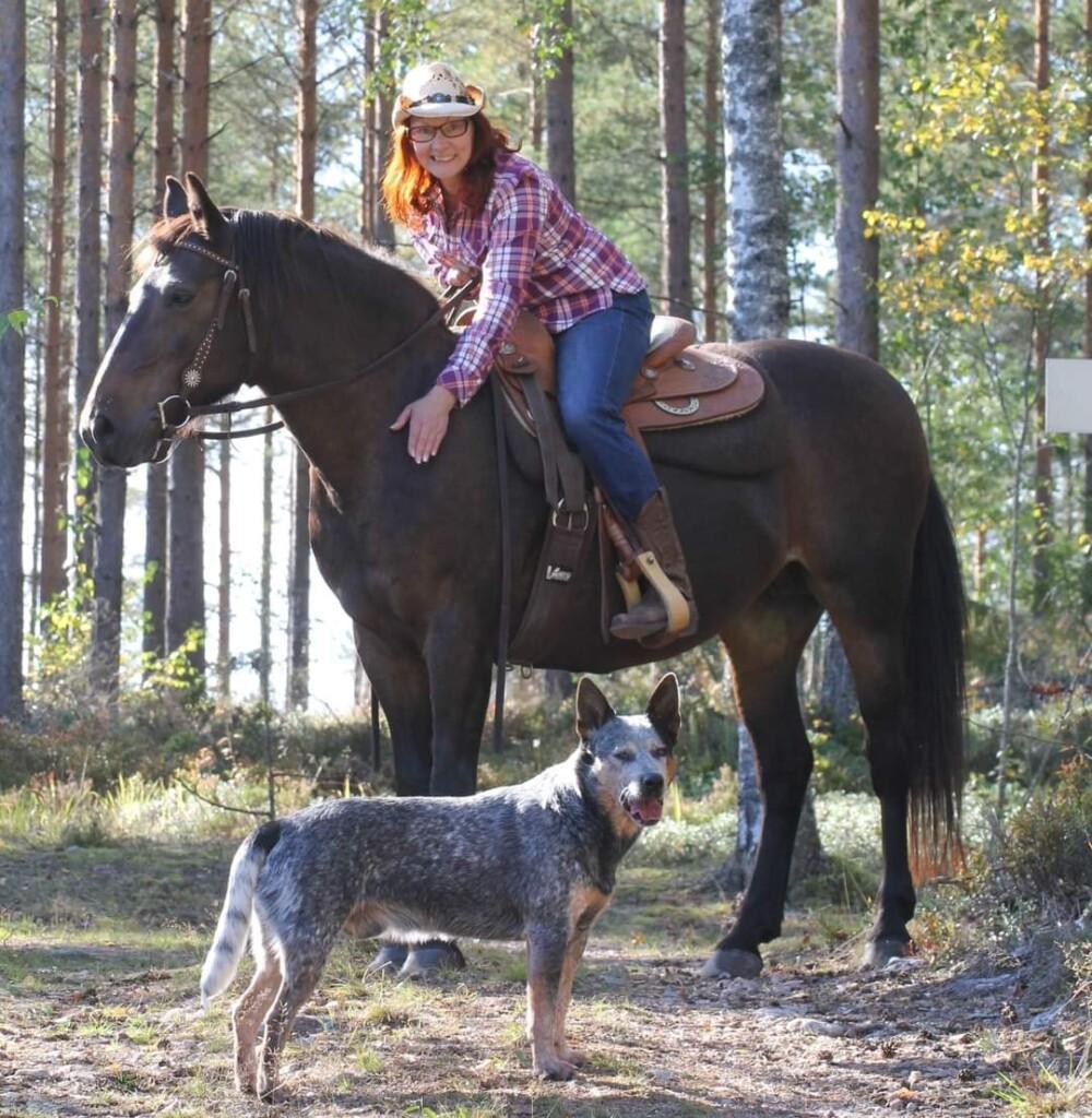 Nainen hevosen selässä cowboy-hattu päässä. Koira seisoo hevosen edessä.