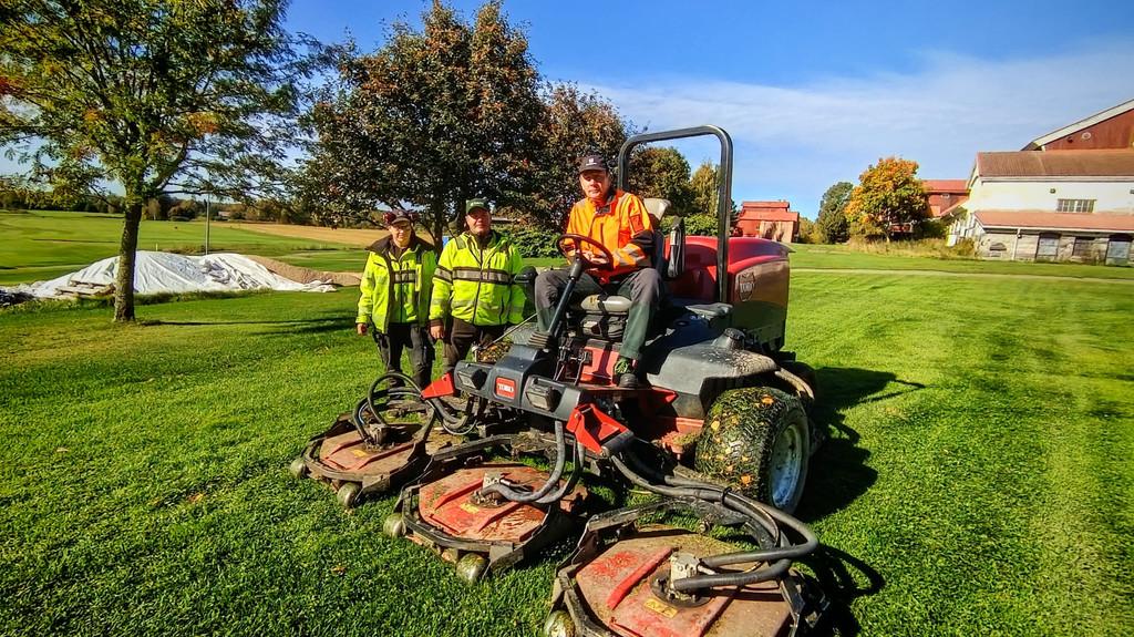 kolme henkilöä golfkentällä. ihmisillä on työvaatteet. yksi ajaa isoa työkonetta.