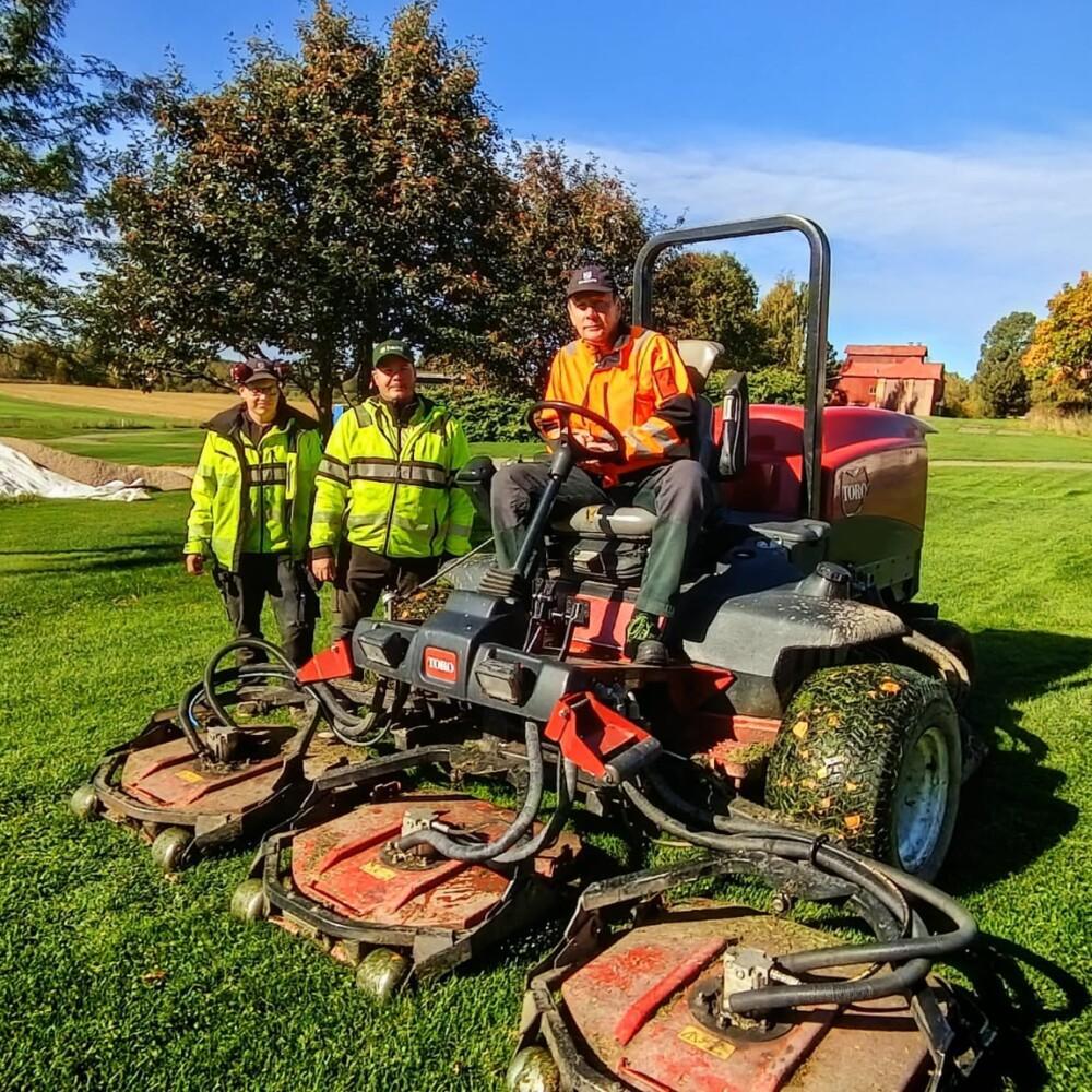 kolme henkilöä golfkentällä. ihmisillä on työvaatteet. yksi ajaa isoa työkonetta.